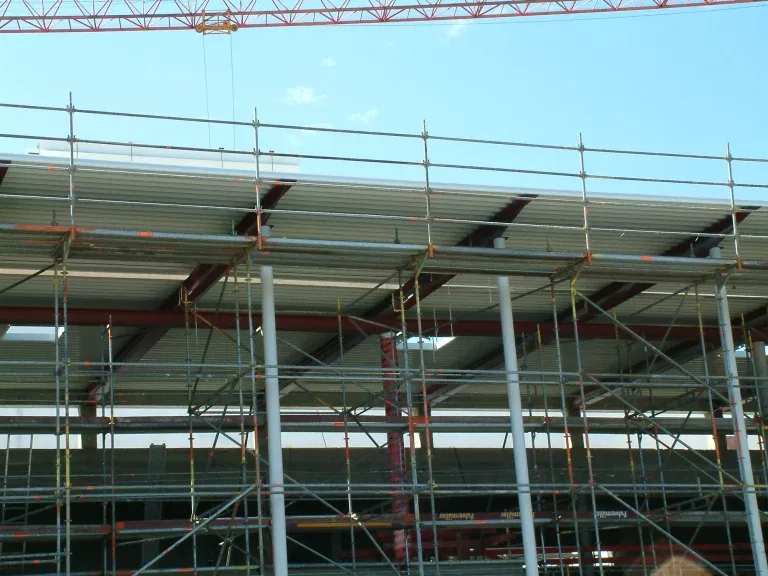 Rohbau eines langgestreckten Multiplexgebäudes mit eingerüsteter Fassade und weit auskragendem Vordach vor blauem Himmel